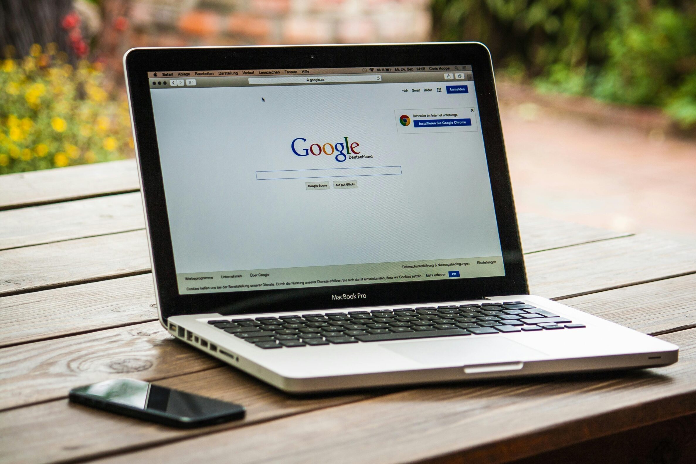 A MacBook Pro displaying Google Search on a wooden table outdoors, next to a smartphone.
